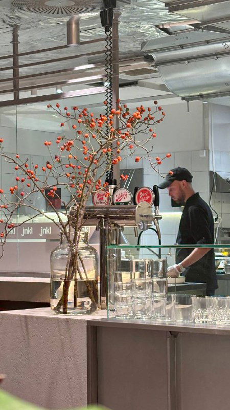 Барная зона и повар за линией раздачи в современном интерьере заведения; на переднем плане высокие вазы с ягодными ветками.