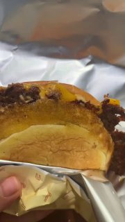 Разрезанный бургер в руке, видно сочную котлету и расплавленный сыр; фольга и упаковка указывают на формат to-go или подачу.