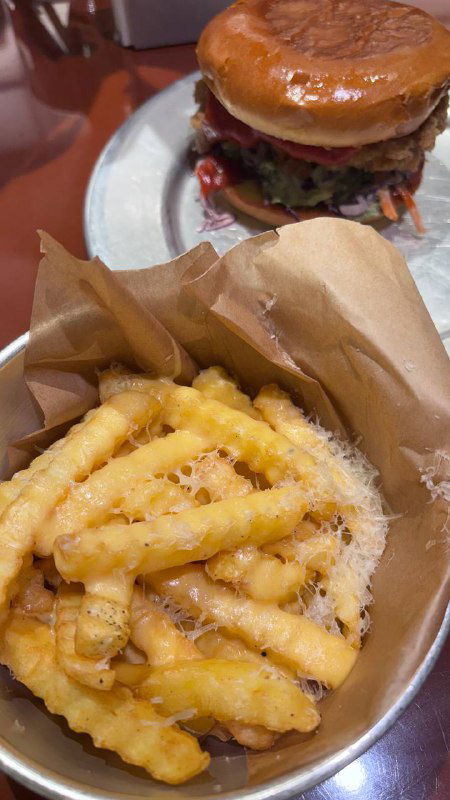 Порция картошки фри в бумажной упаковке на столе рядом с тарелкой и бургером; сверху заметна стружка сыра и текстура картофеля.