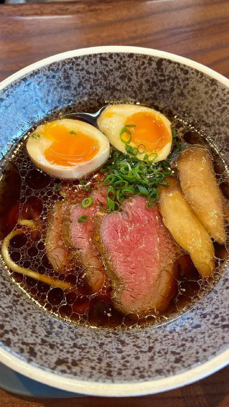 Миска с бульоном и лапшой, два маринованных яйца, ломтики говядины и зелёный лук — тёплое блюдо на столе