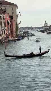Гондола скользит по спокойной воде венецианского канала, на заднем плане видны фасады старых домов и прогуливающиеся люди, типичный городской пейзаж.