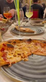 Кусок пиццы на тарелке крупным планом рядом с бокалами аперитивов и тарелками на столе ресторана; уютный кадр итальянского ужина.