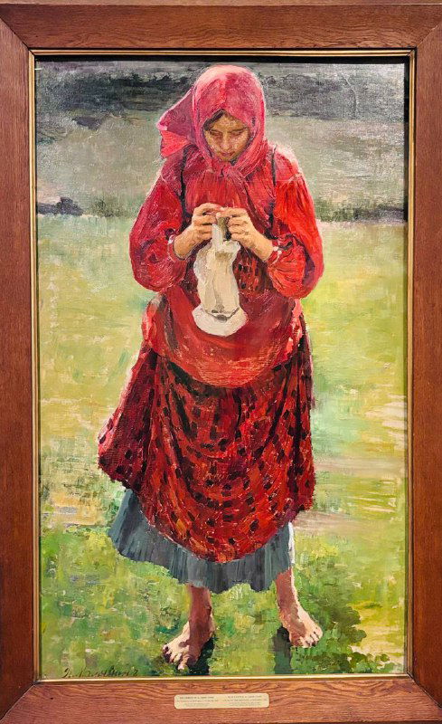 Вертикальная картина сельской женщины в красной одежде, занятой вязанием; спокойная бытовая сцена в контрасте с интенсивностью цвета на фоне.