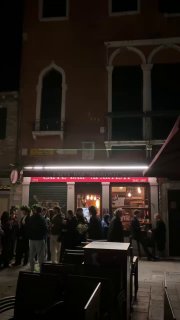 Ночная очередь людей у входа в ресторан или бар в Венеции, подсвеченный фасад и проходящие мимо горожане — сцена вечерней городской жизни.
