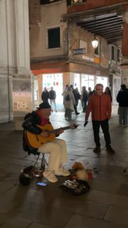 Уличный музыкант в тёплой одежде играет на гитаре на площади, рядом слушают прохожие; атмосферный ночной городской кадр с уличным искусством.