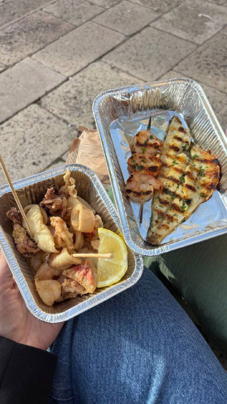 Уличная еда во фольге: чикетти и кусочек рыбы в алюминиевых контейнерах, видно на коленях автора у канала во время быстрой перекуски.