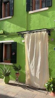 Крупный план зелёного фасада дома с тёмно-зелёными ставнями и белой занавеской, мощёная улочка — бытовая сцена островной Венеции.