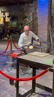 Мастер муранского стеклодува в мастерской за рабочим столом демонстрирует технику выдувания стекла, рядом инструменты и нагретая печь.