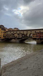 Вид на мост Понте Веккьо через реку Арно: исторические дома на мосту, река и облачное небо — классическая панорама Флоренции.