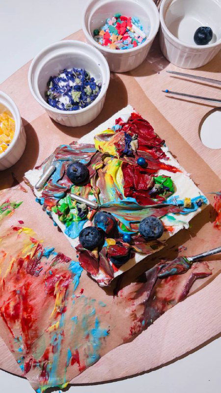 Кусочек торта с яркой разноцветной съедобной росписью, видны посыпки, ягоды и следы кисти на палитре рядом.