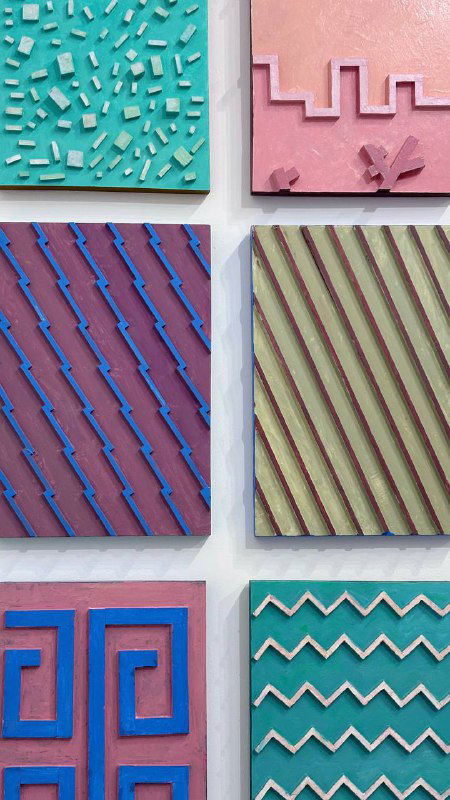 Ряды небольших цветных квадратных панно с геометрическими узорами и пастельными оттенками на белой стене галереи, демонстрация абстрактной серии работ.