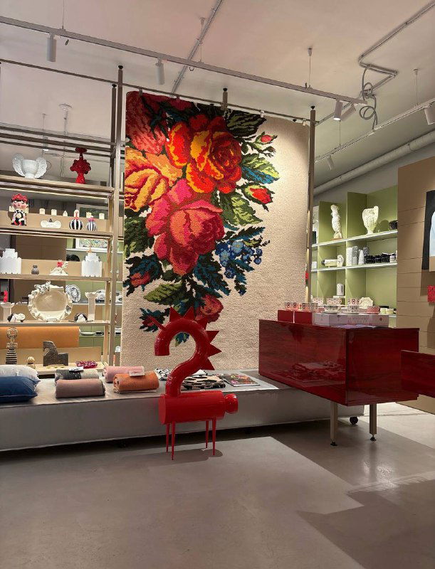 Большой декоративный гобелен с яркими цветами в витрине магазина, на фоне полок с предметами декора и мебелью в стиле концепт‑шопа.
