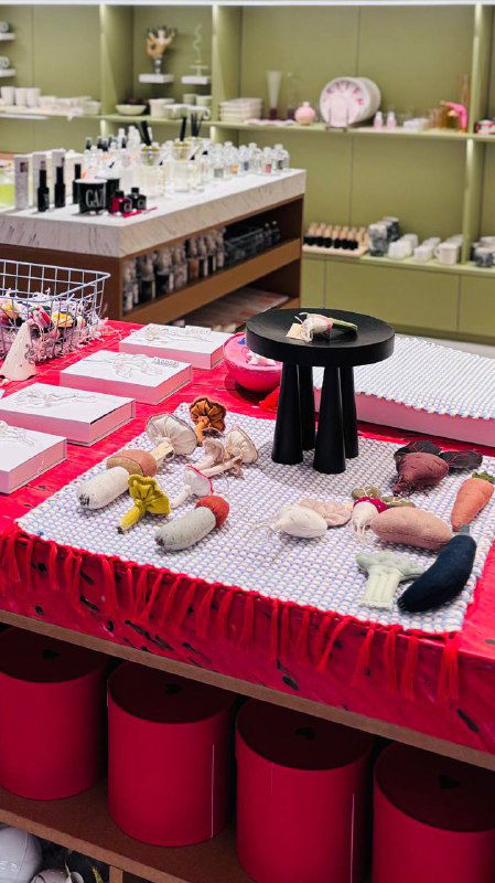 Ярко оформленный стол с текстильными изделиями и мелкими игрушками: на переднем плане мягкие куклы и элементы витрины магазина.