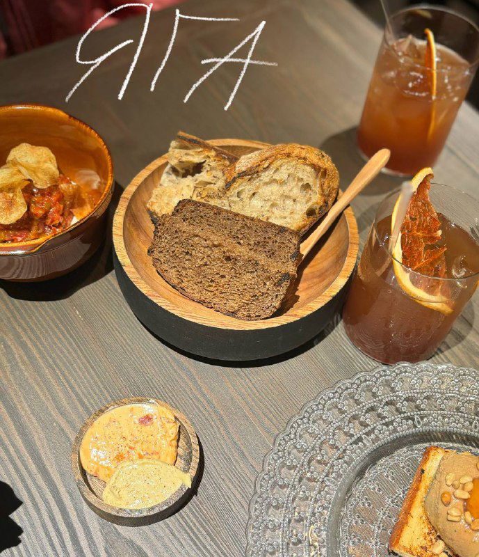 Деревянная миска с разными видами хлеба, бокалы с напитками и закуски на столе; подача и атмосфера, подпись «ЯГА».