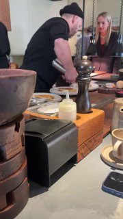 Шеф за барной стойкой в процессе готовки: посуда, соусы, мельница с перцем и наблюдающие гости, атмосферный кадр.