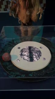 Экспонат с интерактивным столом и круговой панелью — часть инсталляции, рядом виднеется посетитель и тёмный интерьер зала.