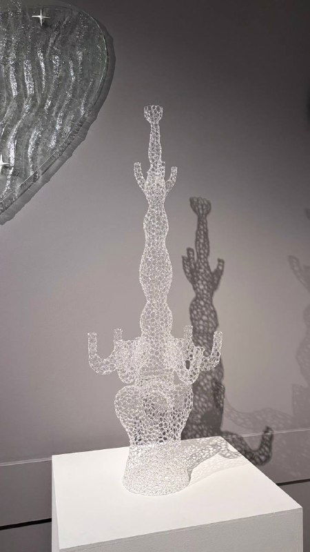 Прозрачная скульптура из точечных стеклянных элементов на пьедестале в галерее Heritage, отбрасывающая сложную тень на белую стену экспозиции.