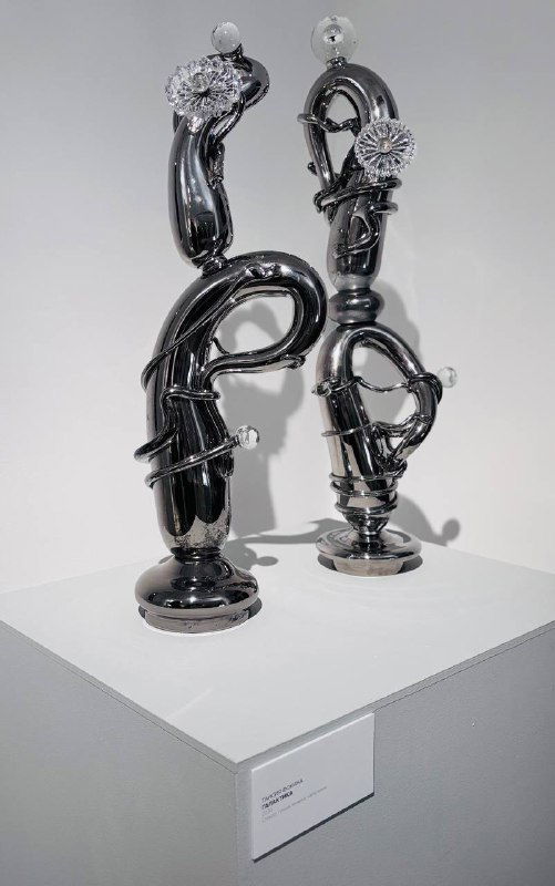 Две высокие глянцевые чёрные стеклянные скульптуры с декоративными элементами на пьедестале в галерее, современная абстрактная работа.