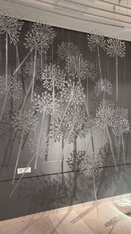 Стена, покрытая множеством тонких стеклянных элементов, напоминающих соцветия одуванчиков, которые создают сложные тени и ритм на задней стене выставки.