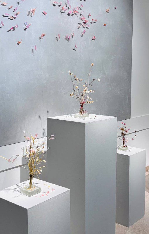 Три небольших композиции на постаментах с миниатюрными стеклянными букетами и разбросанными лепестками в минималистичном музейном интерьере Heritage.