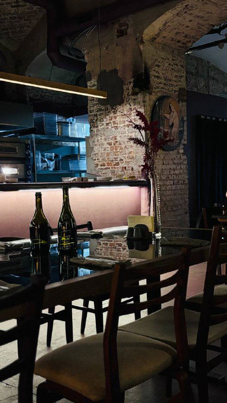 Тёмный зал ресторана с длинным деревянным столом, стульями и открытой кухней на фоне кирпичной стены и bottles вина на полке