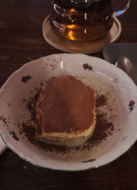 Крупный план порции тирамису на фарфоровой тарелке, посыпанного какао; рядом стеклянный чайник и чашка — десерт в ресторане