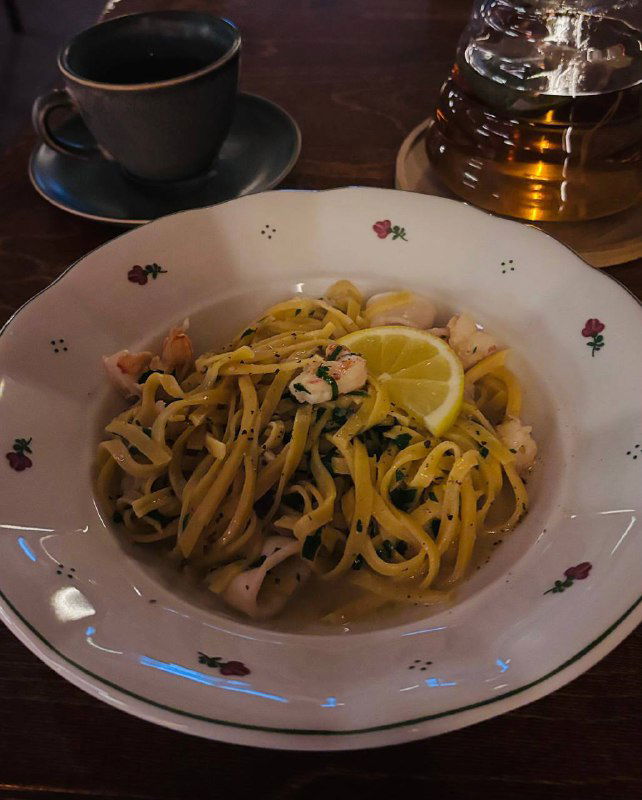 Тарелка пасты с ломтиком лимона и кусочками морепродуктов, сервированная рядом с чашкой чая на деревянном столе в ресторане