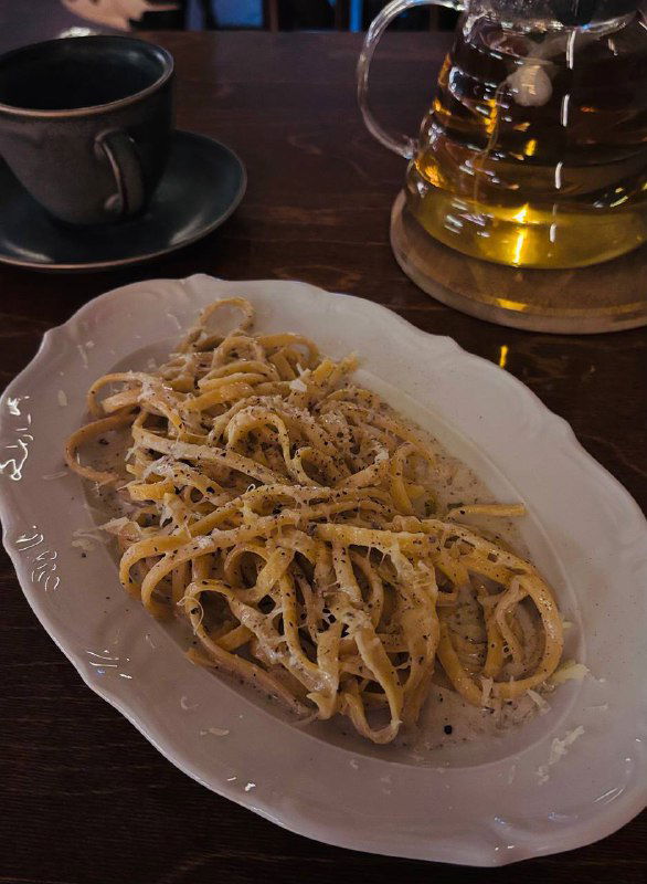 Паста тальятелле, щедро посыпанная тёртым сыром и чёрным перцем в белой овальной тарелке на тёмном столе рядом с чашкой