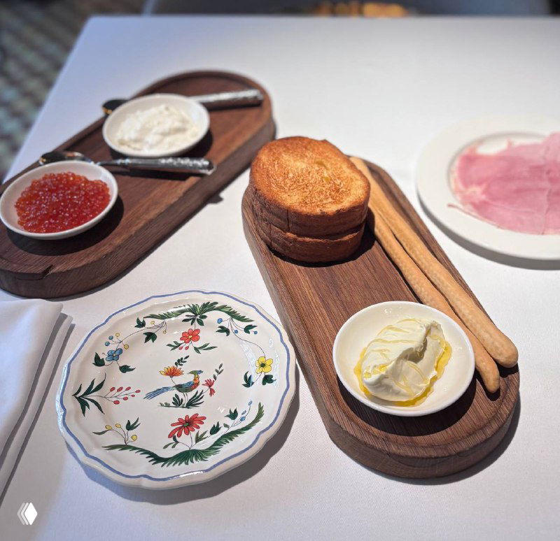 Вид сверху на сервировку: тарелка с икрой, мисочка со страчателлой, бриошь и ломтики прошутто на деревянных досках, белая скатерть.