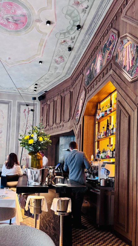 Интерьер ресторана Brera: барная зона с лепниной и витринами, цветы у стойки и посетители, заходящие в зал с высоким декоративным потолком.