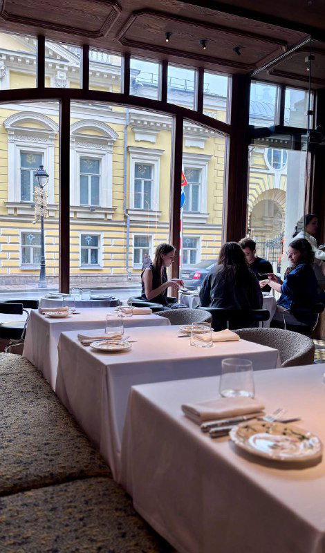 Вид через окно на фасад соседнего здания с классической архитектурой; посетители у оконных столиков завтракают в Brera на Большой Никитской.