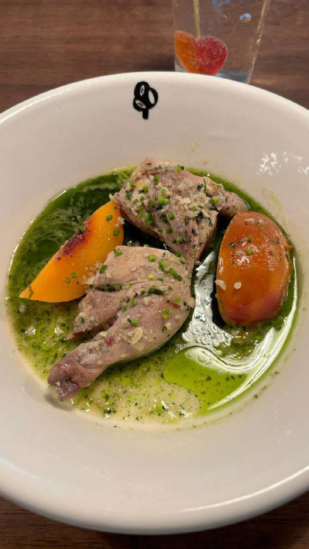 Блюдо с томлёным мясом птицы в белом вине, рядом дольки нектарина и зелёный масляный соус, подано в белой глубокой тарелке на столе.