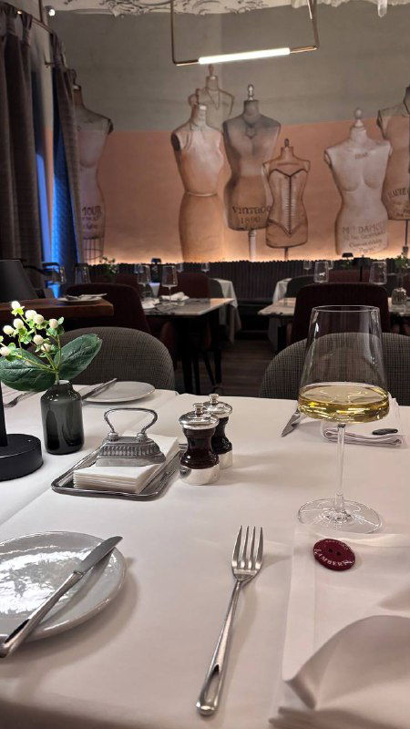 Крупный план сервировки за столом в Sartoria Lamberti: бокал вина, приборы и салфетка на фоне декоративных манекенов и мягкой подсветки интерьера.