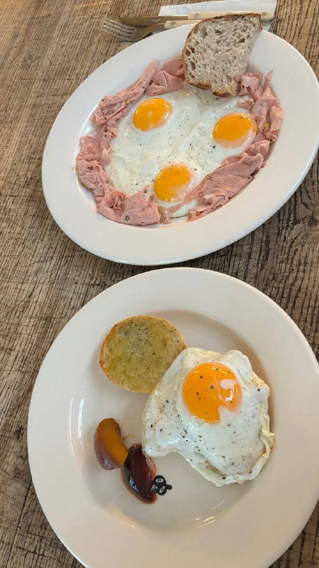 Два блюда на столе: яйца-глазунья и тарелка с гарниром и ломтиком хлеба, на переднем плане видно украшение из мяса и соуса.