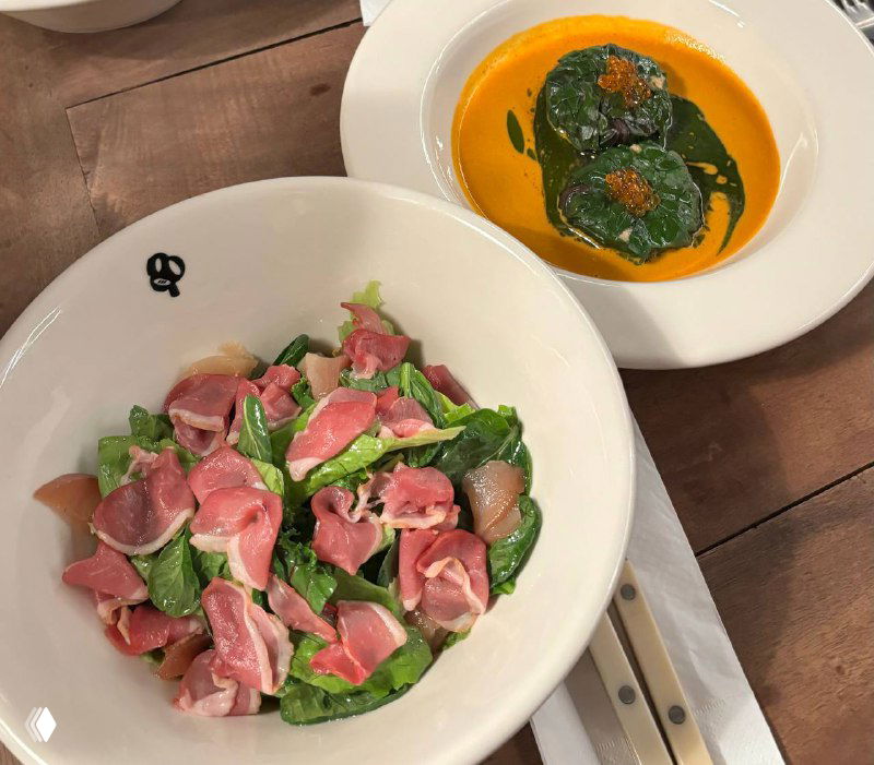 Тарелка с ярко-оранжевым супом и зелёной пастой или пюре, рядом миска со свежим салатом и ломтиками мяса, тёплая подача блюд.