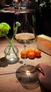 Бокал белого вина на столе рядом с маленькой вазой и мандаринами, отражение света и часть интерьера ресторана на заднем плане.