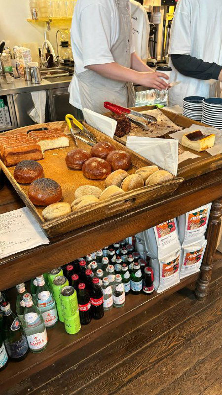 Прилавок с булочками, маффинами и упаковками, видны руки персонала и элементы кассы — типичная сцена в пекарне самообслуживания.