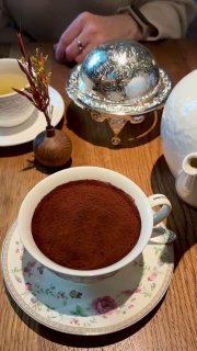 Классический тирамису в холодной тонкой чайной чашке, сверху ровный слой какао; сервировка на столе ресторана Rebecca с серебристой крышкой рядом.