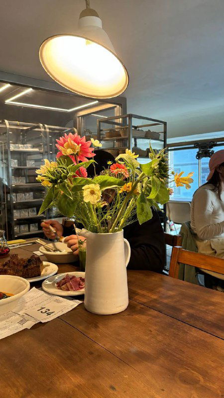 Ваза с цветами стоит на деревянном столе в интерьере пекарни, вокруг гости и спокойная уютная атмосфера заведения.