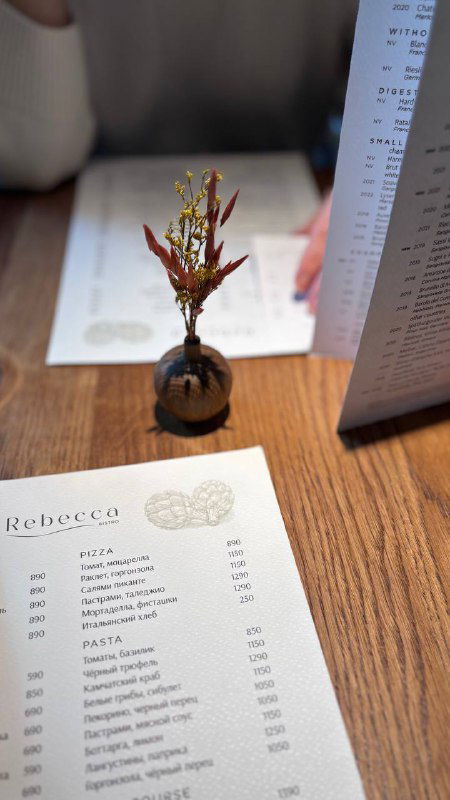 Маленькая керамическая ваза с веткой рядом с меню на деревянном столе — деталь сервировки и эстетики в Rebecca.