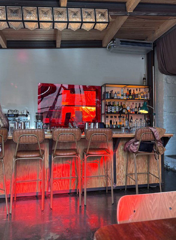 Барная стойка с деревянными стульями и яркой красной подсветкой, пол из шлифованного бетона и стеллаж с бутылками на стене заведения.