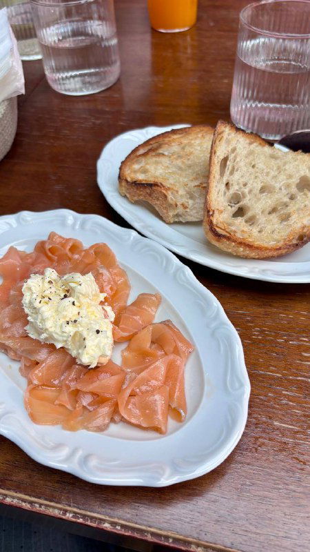 Подача лосося с творожной намазкой и кусочками хлеба на тарелке, видны столовые приборы и ресторанная обстановка.