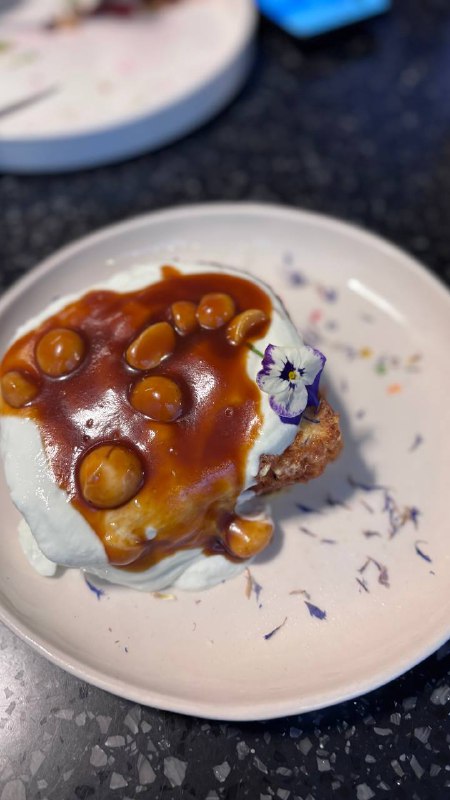 Френч-тост с фиалковой рикоттой, лимонным соусом и карамелизованной макадамией на белой тарелке, украшенный съедобными цветами.