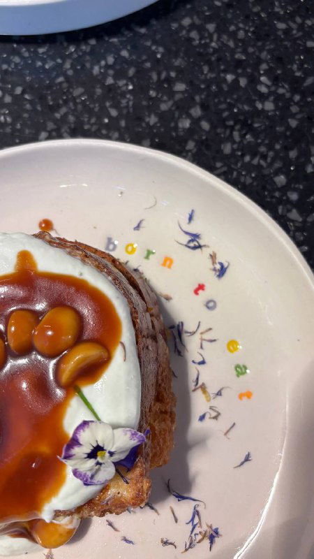 Крупный план френч-тоста с карамелизованной макадамией и фиалковой рикоттой, смещённый кадр, видны съедобные цветы на тарелке.