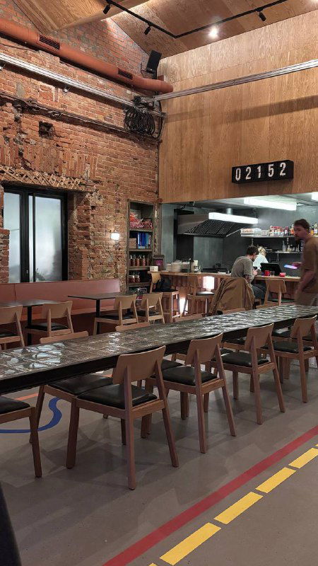 Общий ракурс зала бара с длинным стеклоблоковым столом и рядами стульев, кирпичная отделка стен и открытая кухня на заднем плане.