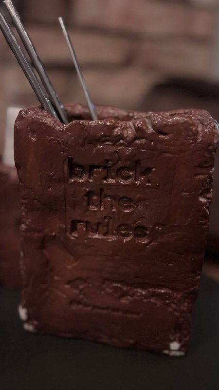 Десерт в форме кирпича с оттиском «brick the rules», крупный план шоколадной глазури и фактуры десерта на тёмном фоне.