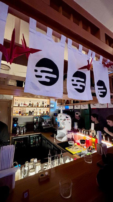 Барная стойка под японскими норен с символикой, на тыльном плане бутылки и барные аксессуары, заметны бумажные журавли.