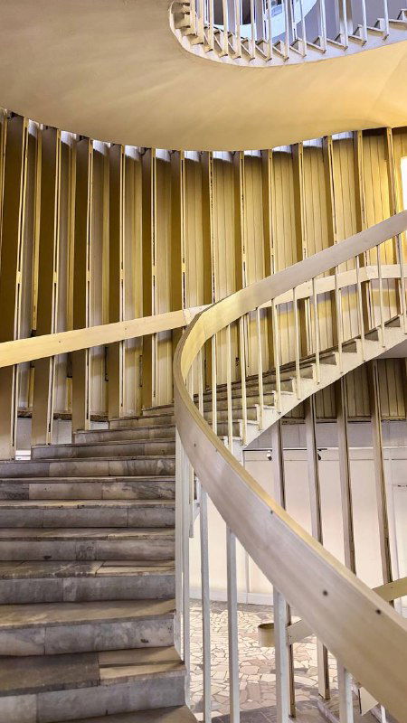 Диагональный ракурс спиральной лестницы: деревянные перила и ступени, повторяющаяся ритмика и мягкая золотистая подсветка в интерьере библиотеки.