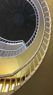 Фрагмент спиральной лестницы с изгибом ступеней и перил, акцент на переходах света и тени по кривой поверхности внутри библиотечного интерьера.