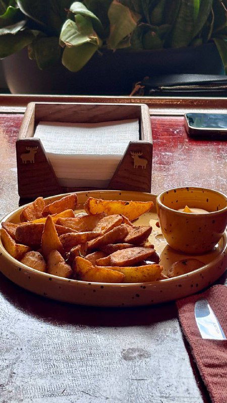 Порция картошечки фри на керамической тарелке с маленькой соусницей на деревянном столе в кафе Mission, рядом салфетки и стерильно простая подача.